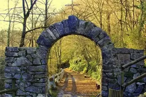 stone arch with small metal cross on top leading into a wooded area