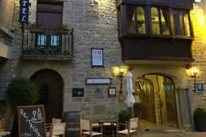 A stone building with a medieval balcony over the entrance. Chairs a table a parasol and a barrel just outside