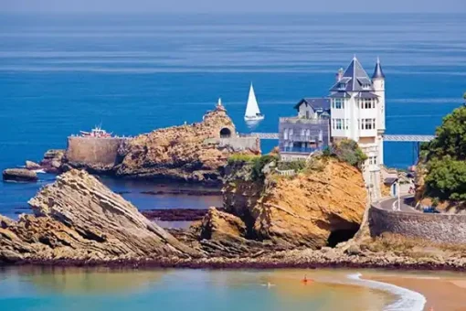 a tall narrow white house among rocks on the coast a bit of beach in the foreground and a sail boat on the sea
